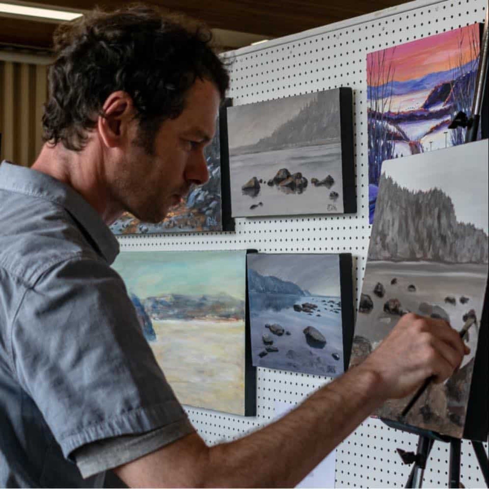Jean Delisle peint une toile de paysage côtier pendant une exposition, d'autres toiles accrochées autour