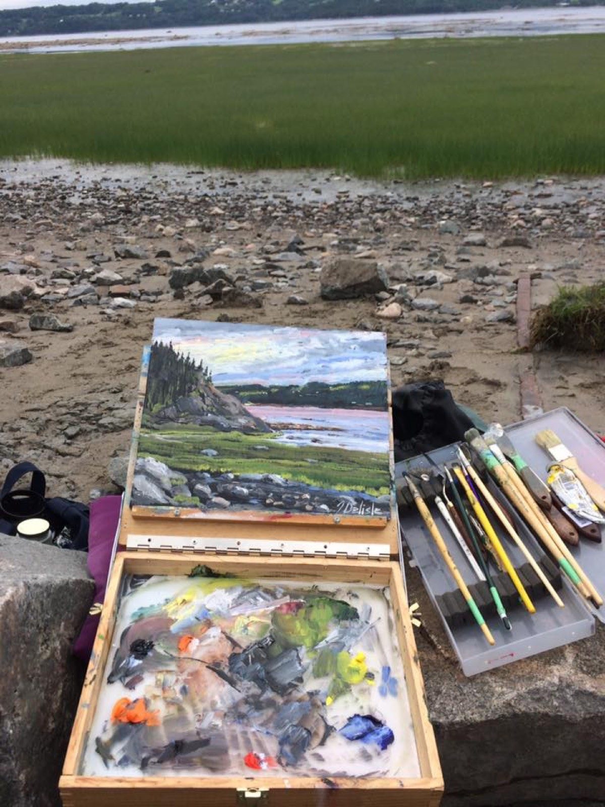 Boîte de peinture ouverte sur la grève, palette et pinceaux, toile du fjord en cours